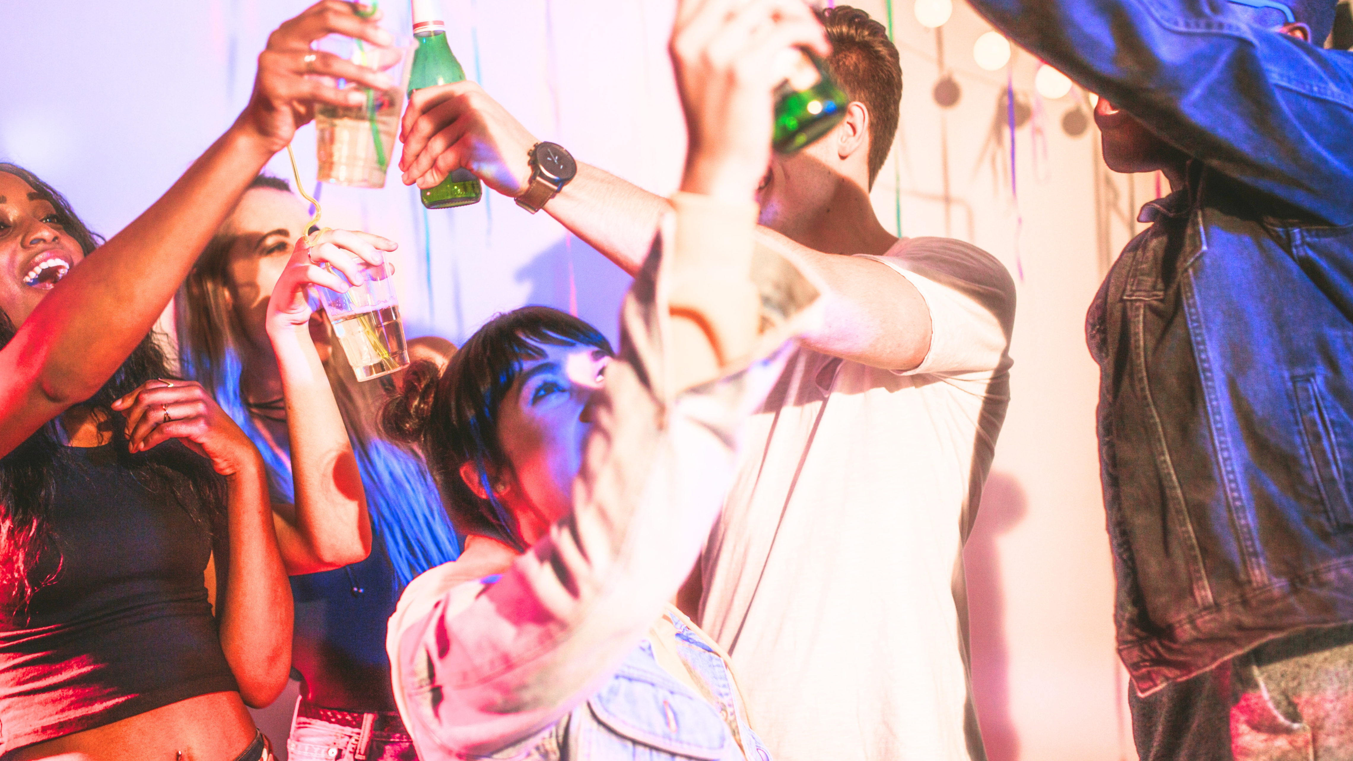 Crowded apartment party mid-night, people raising drinks at a 1st house astrology party, colored lights, packed room, nobody looking at the camera.