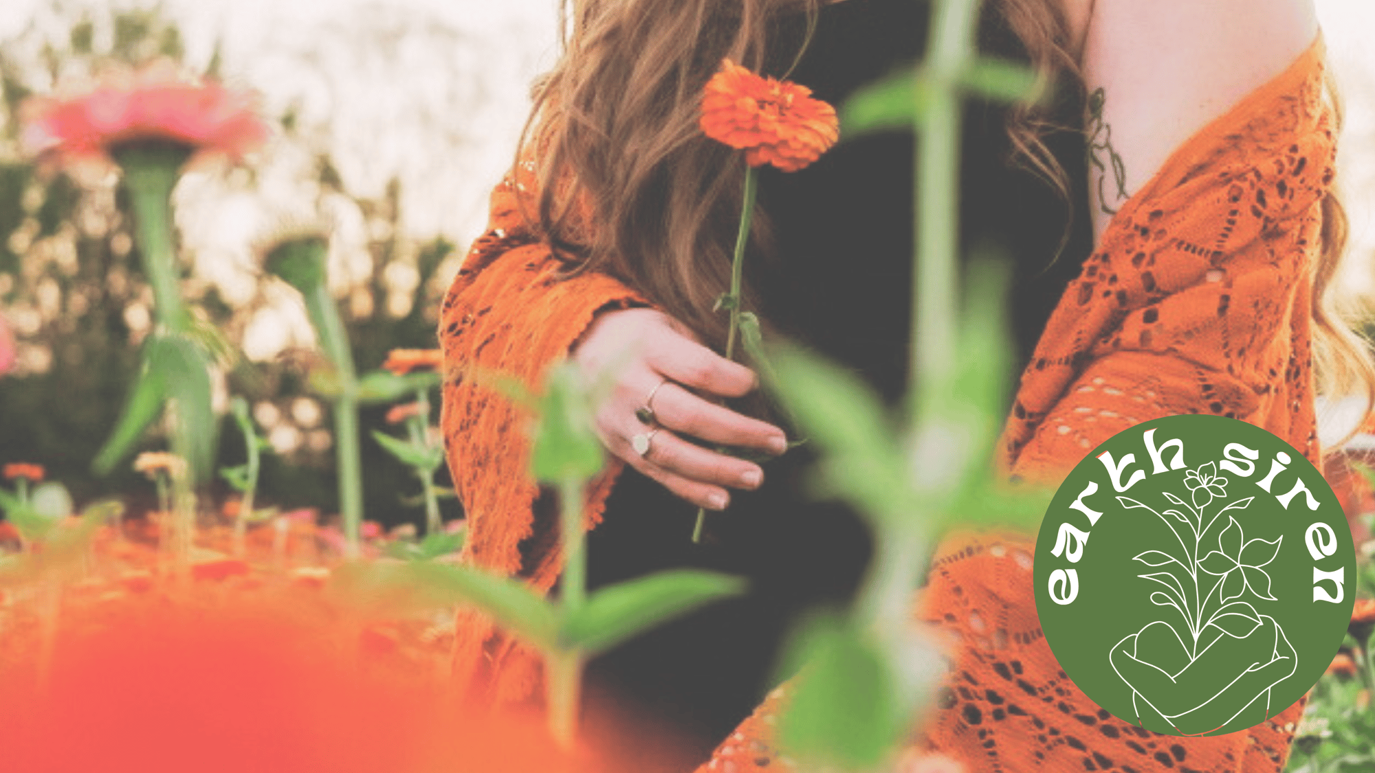 Elizabeth Schirk holding an orange flower in a blooming field, wearing a burnt orange shawl, with the Earth Siren logo in the corner Elizabeth Schirk holding an orange flower in a blooming field, wearing a burnt orange shawl, with the Earth Siren logo in the corner