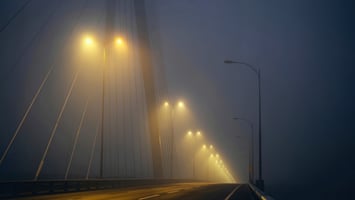 Empty bridge at night fading into dense fog, with evenly spaced streetlights receding into low visibility and no people or vehicles present.
