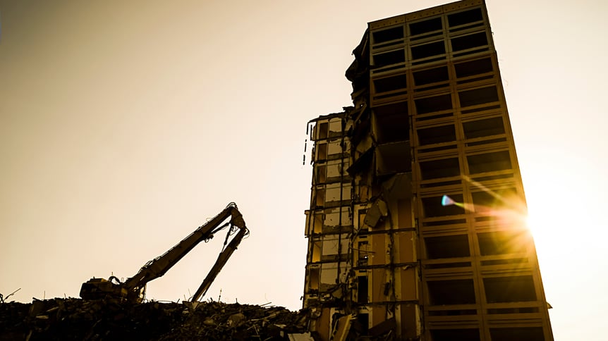 Excavator demolishing a tall building at sunset, with sunlight cutting through the exposed concrete structure