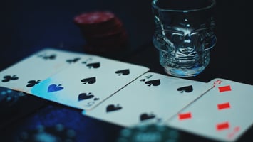 Poker cards and casino chips arranged on a dark table beside a skull-shaped shot glass, illuminated by moody blue lighting, creating a secretive speakeasy atmosphere associated with risk, power, hidden debts, and transformation.