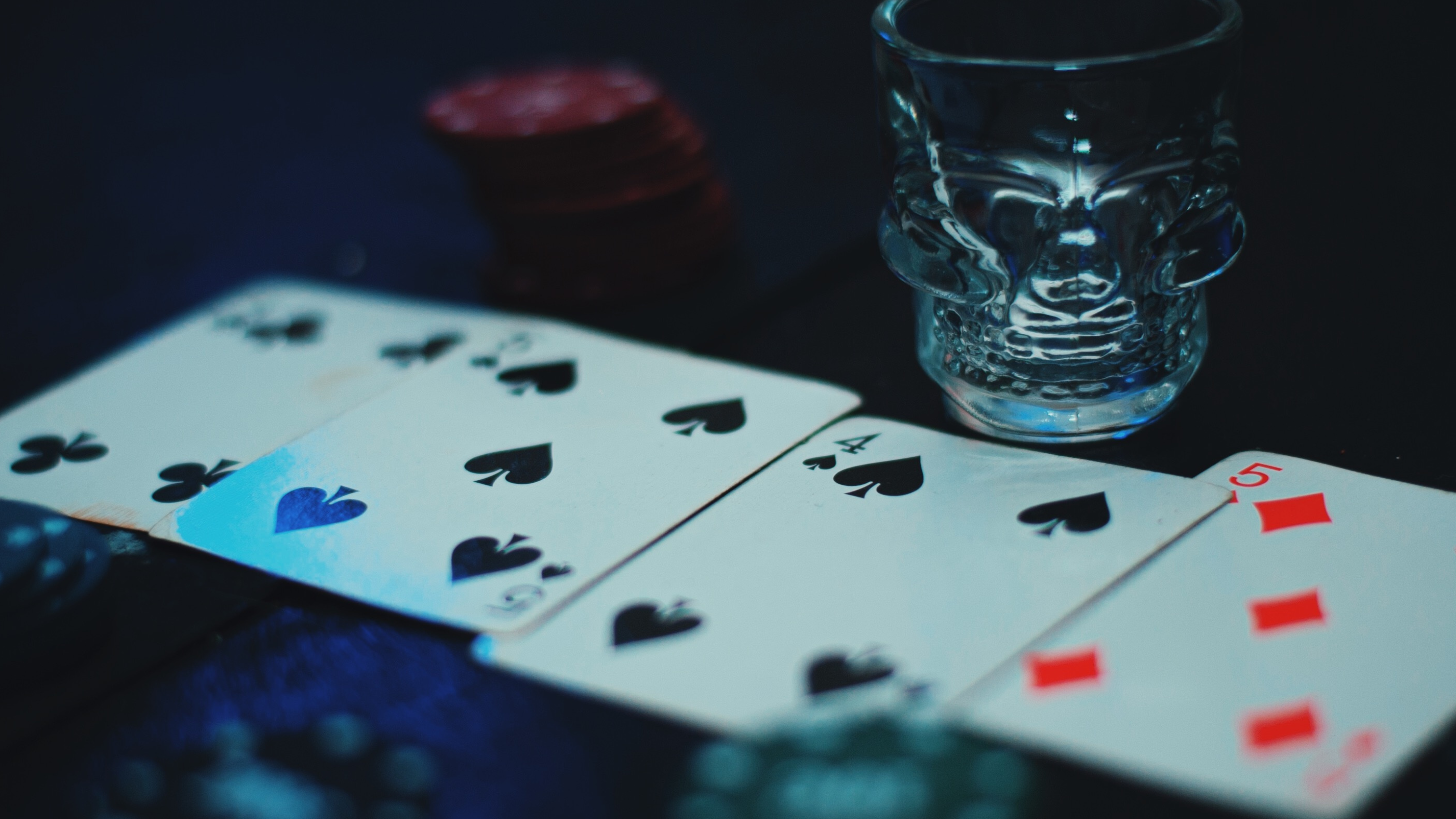 Poker cards and casino chips arranged on a dark table beside a skull-shaped shot glass, illuminated by moody blue lighting, creating a secretive speakeasy atmosphere associated with risk, power, h