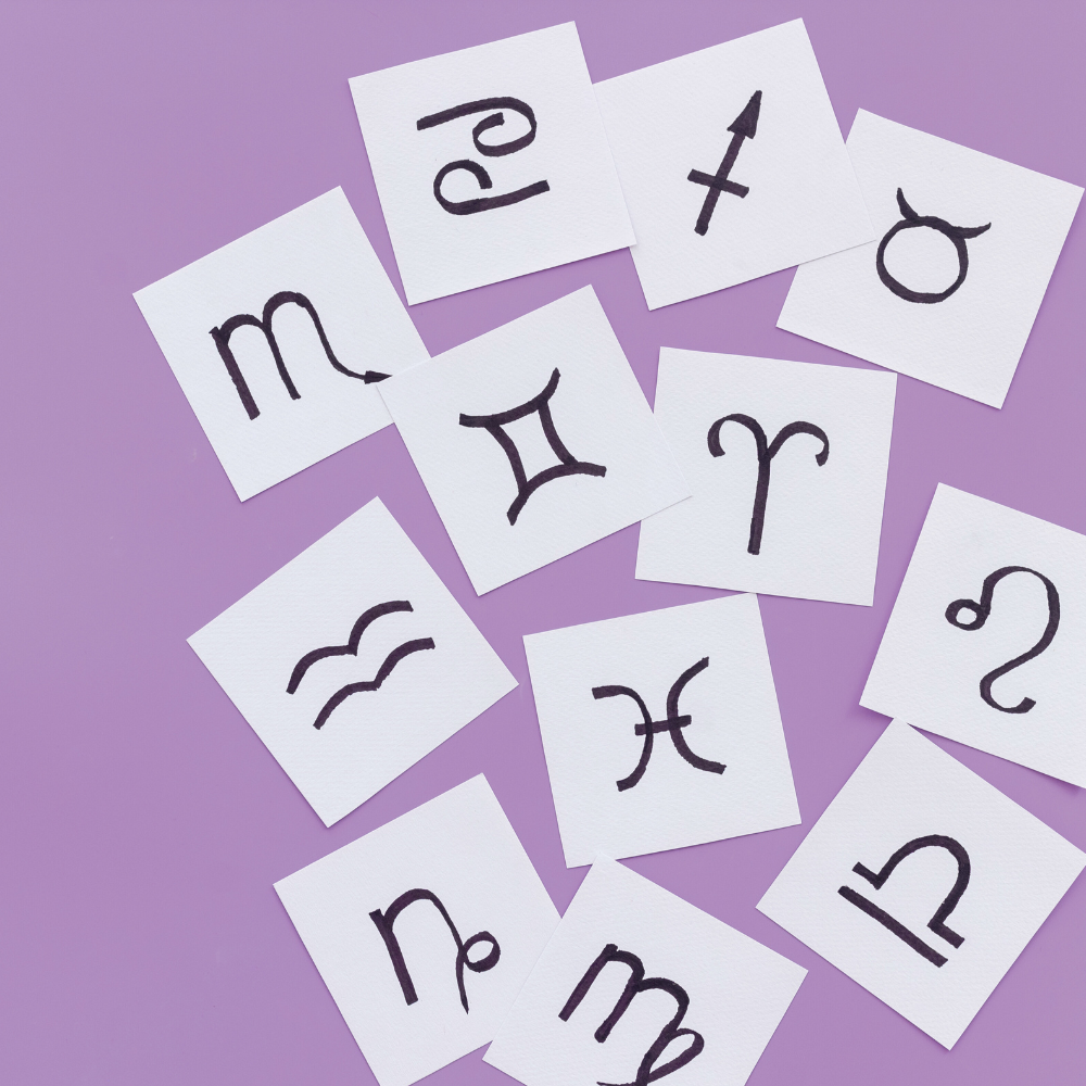 Handwritten zodiac symbols on small paper squares scattered across a purple background, representing astrology and birth chart signs