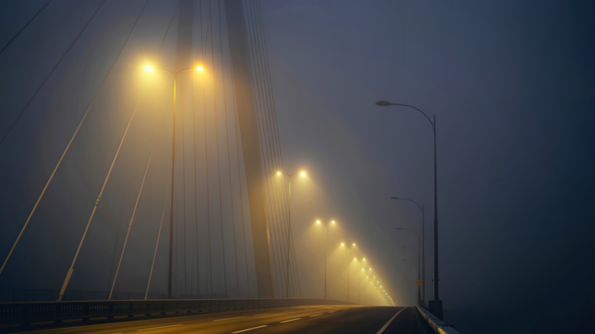 Empty bridge at night fading into dense fog, with evenly spaced streetlights receding into low visibility and no people or vehicles present.